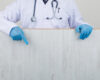 Doctor Showing Wooden Board In White Coat, Gloves Front View.